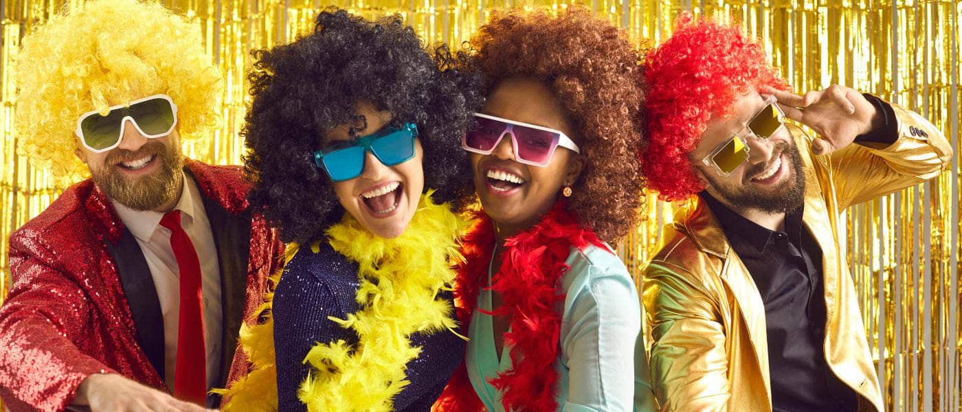 Lustige Fotobox-Momente mit Verkleidungen und Accessoires Vier verkleidete Personen posieren mit bunten Perücken und Sonnenbrillen vor einem goldenen Fotobox-Hintergrund.