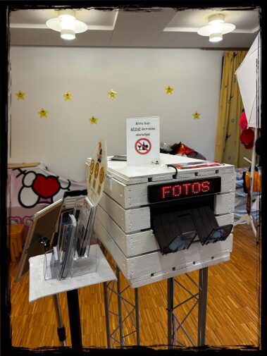 Eine Fotobox auf einer Veranstaltung mit einem "Fotos"-Display und einem Hinweis, dass keine Getränke abgestellt werden dürfen. Die Fotobox ist stilvoll im Holzdesign gehalten, ideal für kreative Event-Erlebnisse. Perfekt für Hochzeiten, Geburtstagsfeiern oder Firmenevents, bei denen Gäste Spaß haben und Erinnerungen festhalten können.