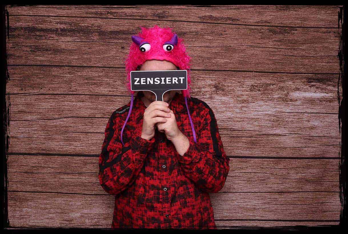 Person mit pinker Monster-Mütze hält ein „ZENSIERT“-Schild aus 3D-Druck vor das Gesicht