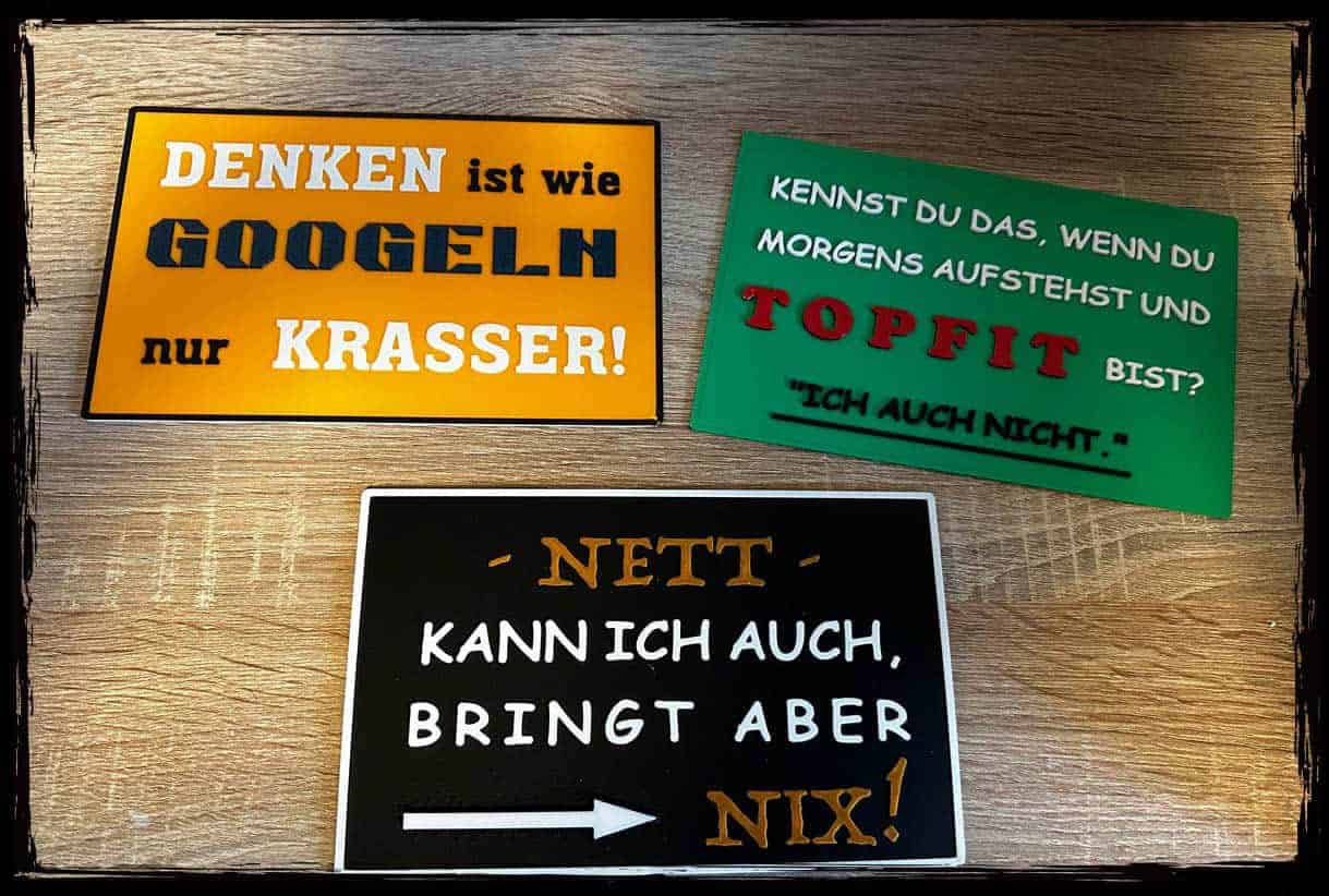 Drei 3D-gedruckte Schilder mit humorvollen Sprüchen in verschiedenen Farben auf Holzhintergrund