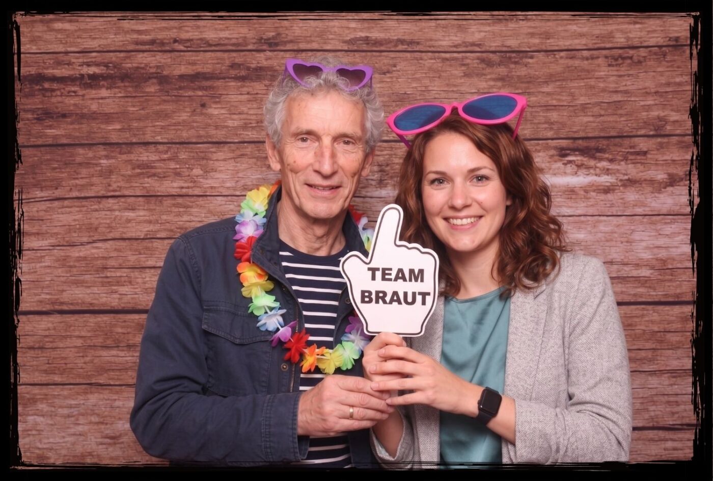 Zwei Personen halten ein Team Braut Fotobox Schild vor einem Holz-Hintergrund