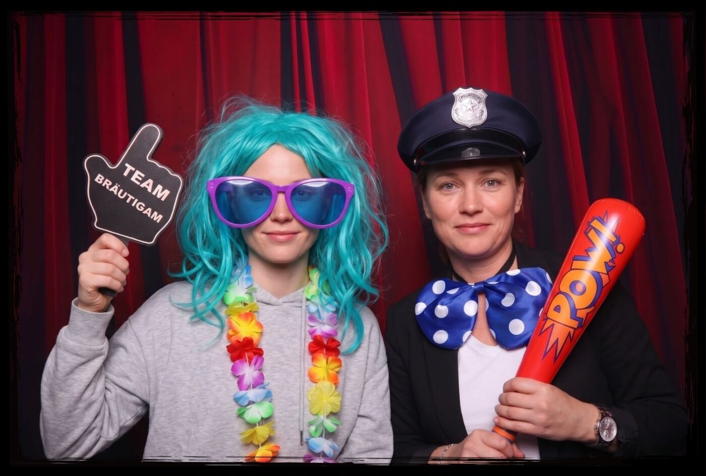 Zwei Personen mit Perücken und Accessoires präsentieren ein Team Bräutigam Schild