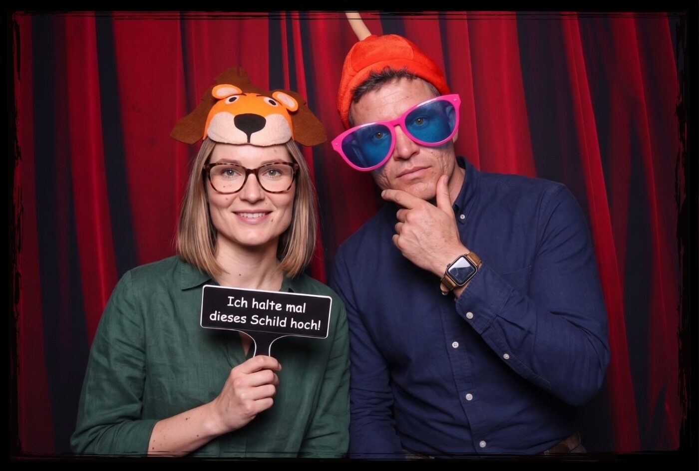 Lustige Fotobox Schilder Requisiten kaufen Frau mit Loewe-Maske und Mann mit bunter Brille halten ein Fotobox Schild hoch