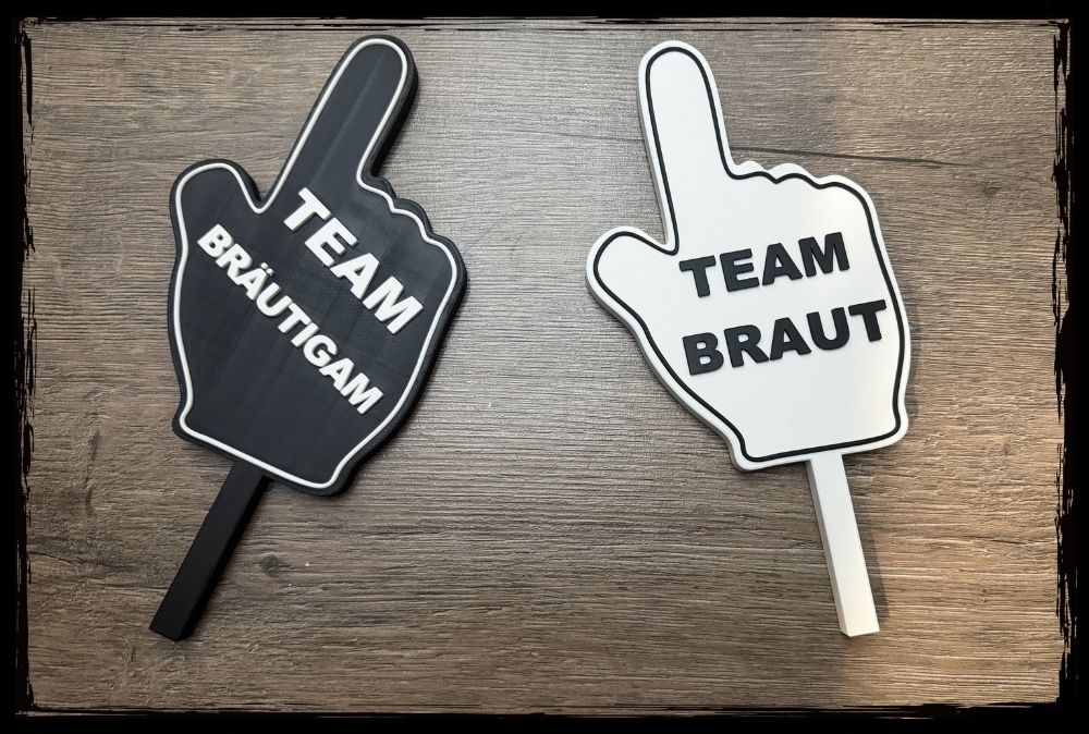 Zwei handförmige Fotobox Schilder mit der Aufschrift Team Braut und Team Bräutigam auf einem Holzhintergrund.