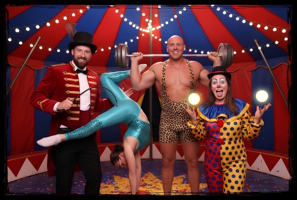 KI Fotobox Sachsen für kreative Zirkusmotive auf deinem Event Vier Personen posieren als Zirkusdirektor Akrobatin Gewichtheber und Clown in einem Zirkuszelt erstellt mit einer KI Fotobox Sachsen
