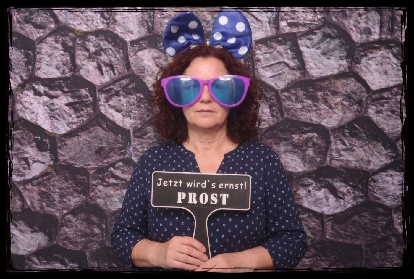 Eine Frau mit riesiger lila Brille und blauer Schleife hält ein Fotobox Props 18. Geburtstag Schild mit dem Text Jetzt wirds ernst PROST