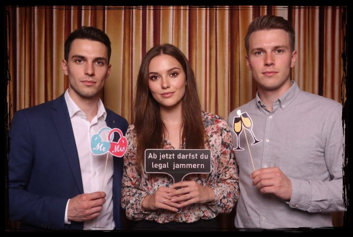 Drei junge Erwachsene posieren mit Fotobox Props 18. Geburtstag wie Sektglaesern und einem Schild mit dem Text Ab jetzt darfst du legal jammern