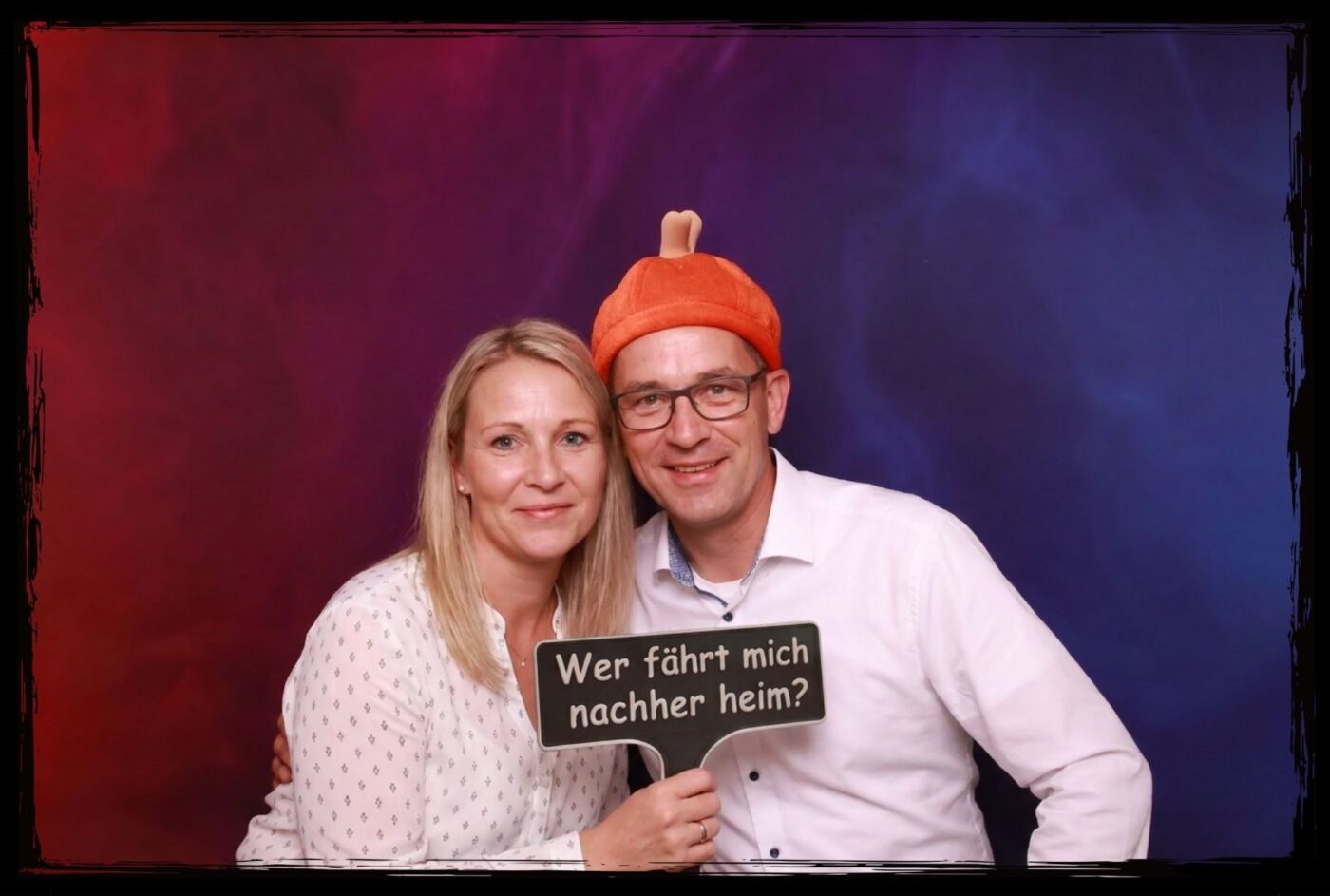 Ein lachendes Paar nutzt Fotobox Props 18. Geburtstag inklusive eines Spruchschildes mit Wer faehrt mich nachher heim und einer Partyhut-Requisite
