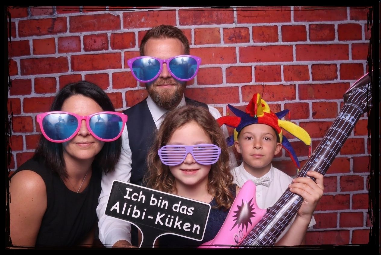 familie mit zwei kindern posiert vor einer backsteinwand mit bunten brillen narrenkappe aufblasbarer gitarre und einem schild mit dem aufdruck ich bin das alibi-küken