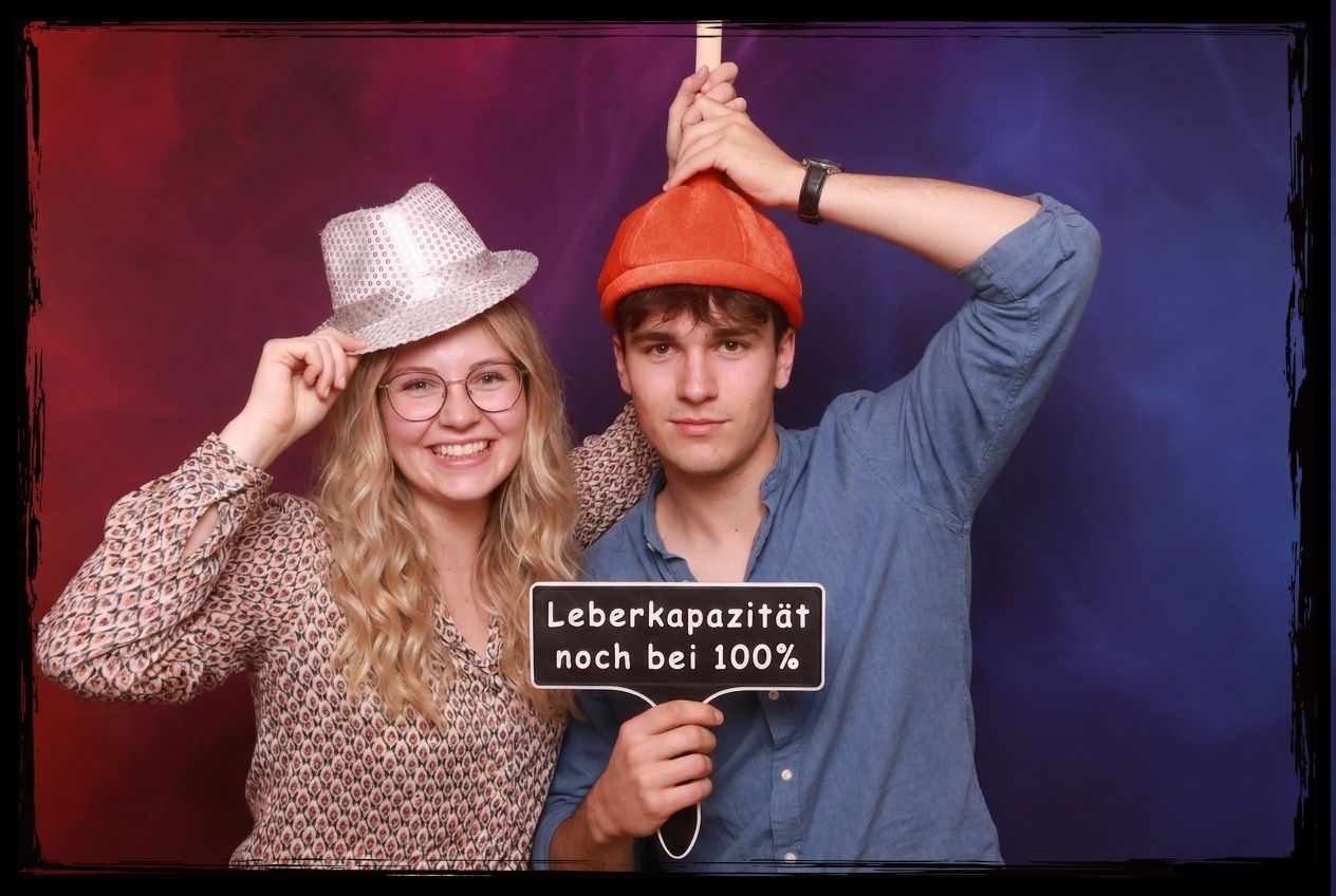 junges paar in einer fotobox mit witzigen hueten hält ein schild mit dem text leberkapazität noch bei 100 prozent