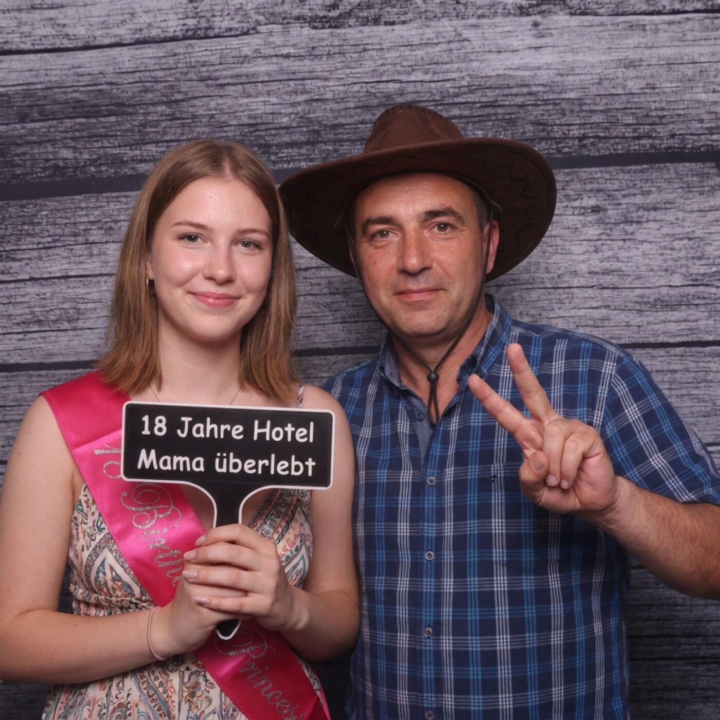 Junges Mädchen mit Geburtstagsschärpe und ein älterer Mann mit Cowboyhut posieren mit dem Hotel Mama Schild