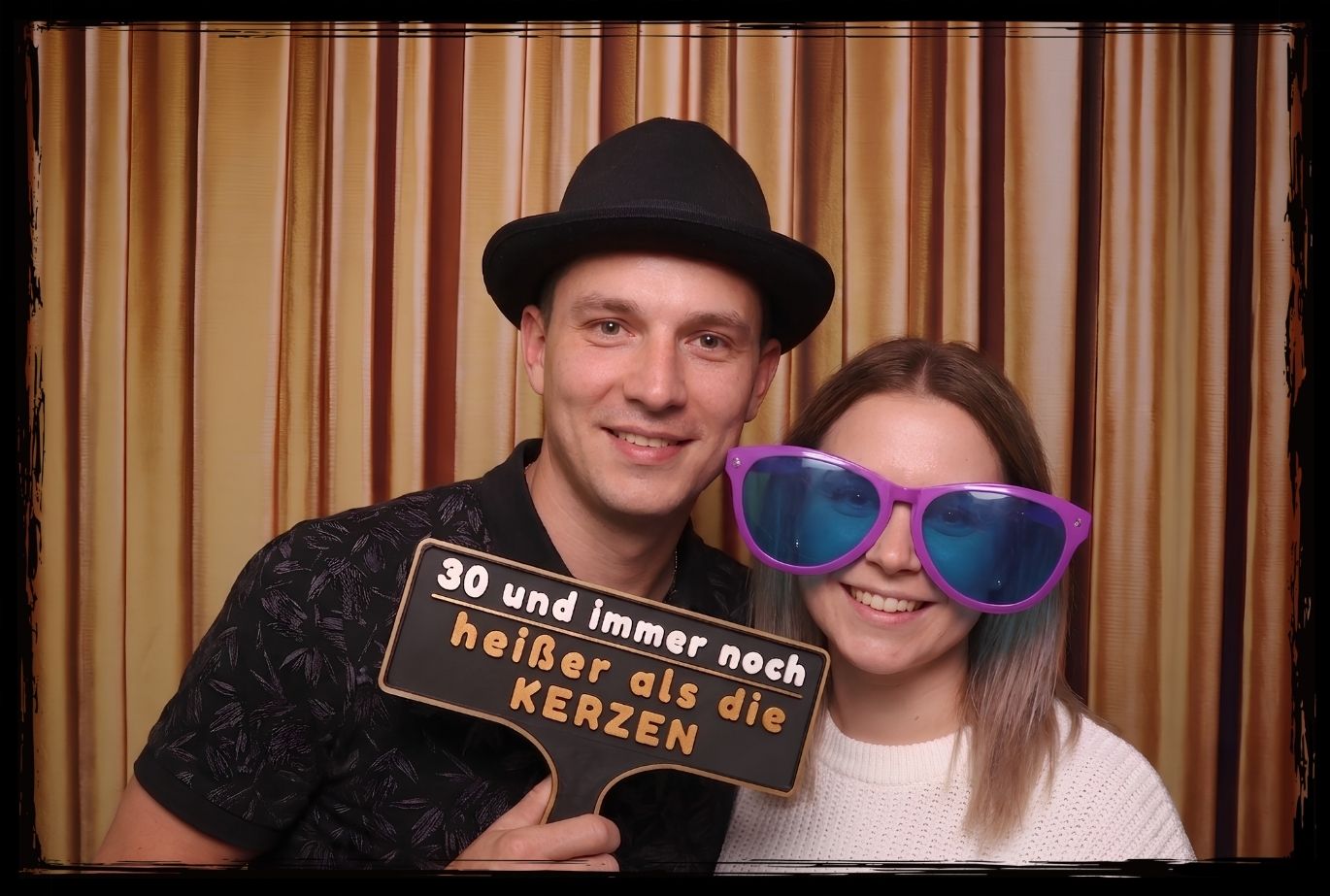 Mann und Frau vor einem goldenen Vorhang nutzen Fotobox Schilder für den 30. Geburtstag, der Mann präsentiert den Spruch 30 und immer noch heißer als die Kerzen