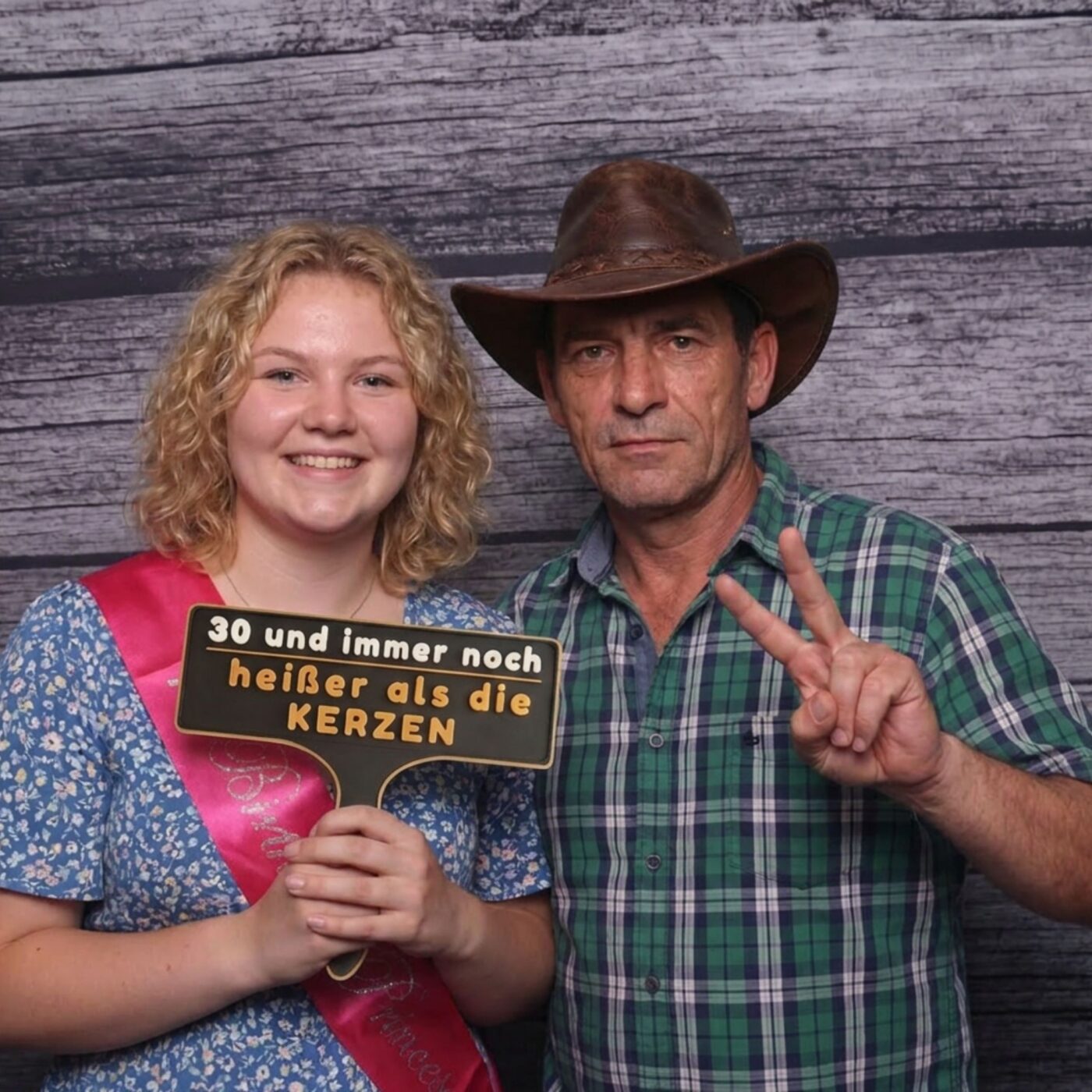 Madchen mit Geburtstagsschaerpe und Mann mit Cowboyhut posieren mit Schild vor Holzwand